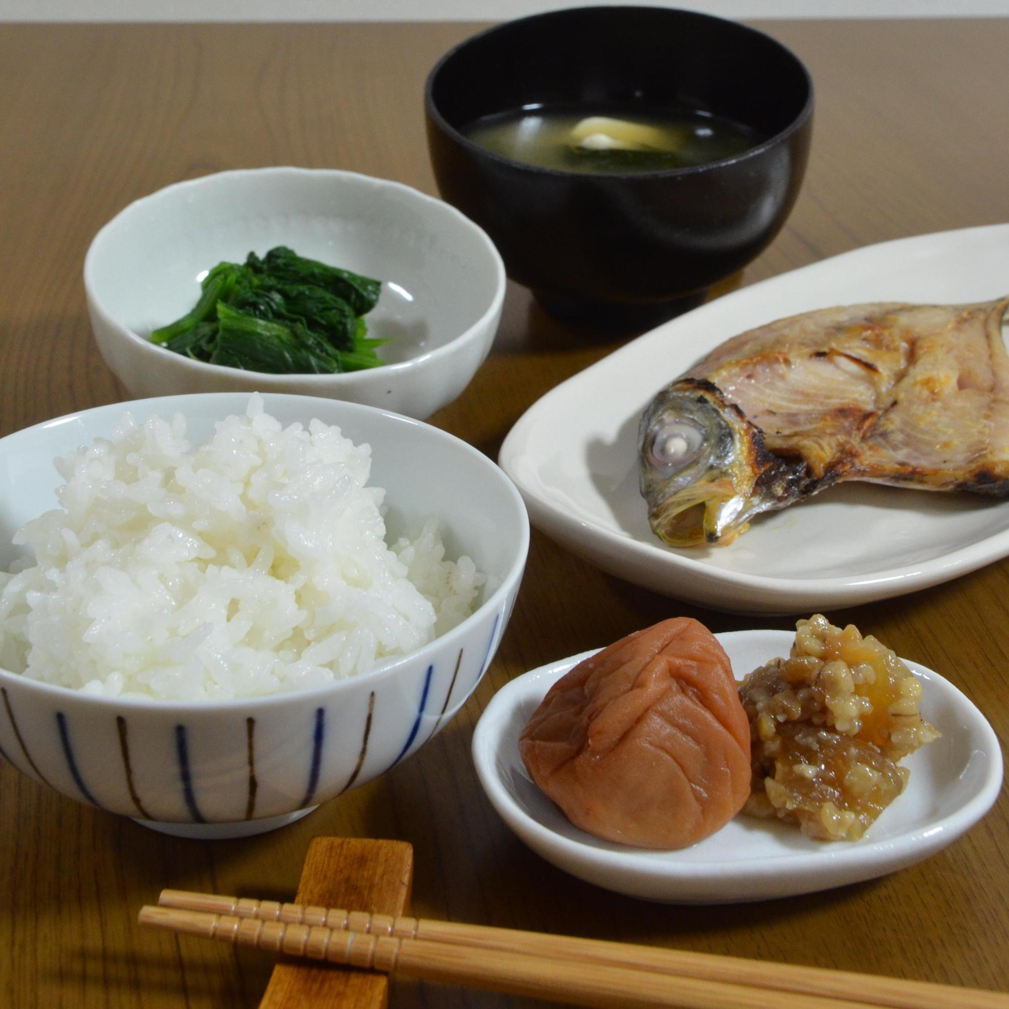 ごはん、みそ汁、焼き魚、漬物、小鉢がテーブルに並ぶ写真